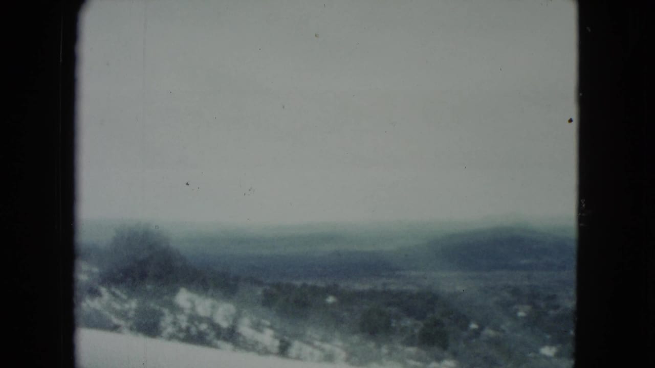 Muted 8mm view of Nevada hills and pale ground patches, partially framed by a dark car-window edge.
