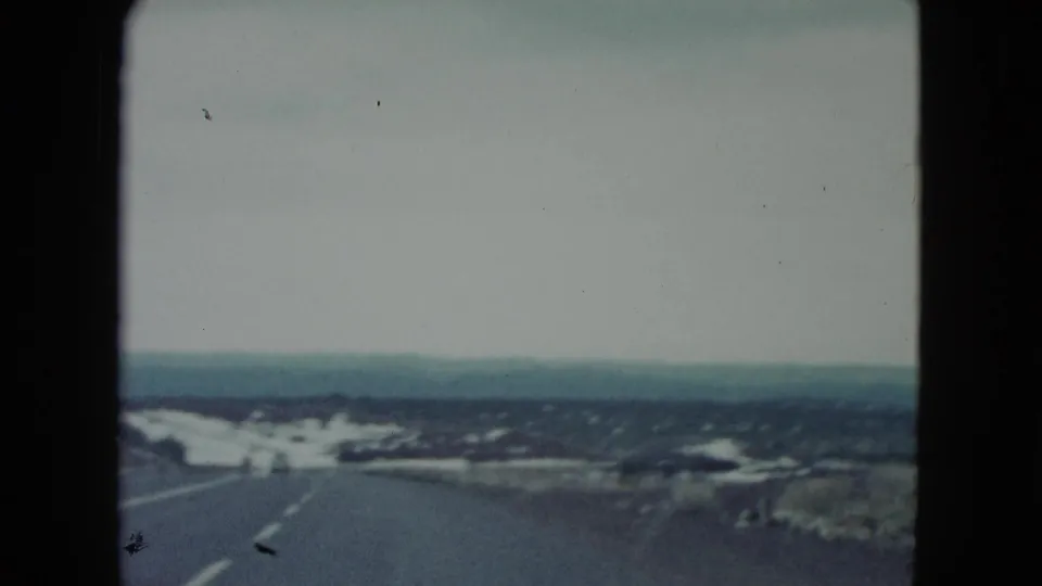 1960 Nevada roadside view from a moving car, with road edge, sparse terrain, pale sky, and distant low hills.