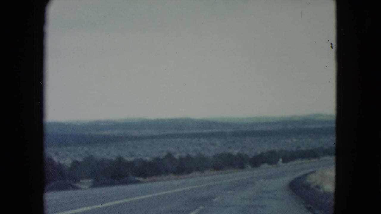 1960 Nevada 8mm travel view of a winding desert road, sparse shrubs, pale ground cover, and distant hills beneath an overcast sky.
