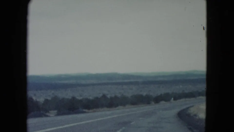 1960 Nevada 8mm travel view of a winding desert road, sparse shrubs, pale ground cover, and distant hills beneath an overcast sky.