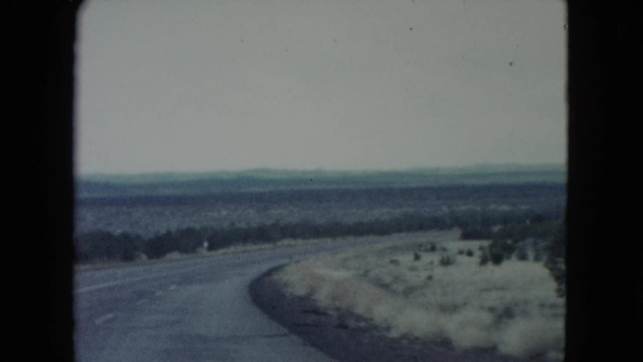 1960 Nevada home-movie frame showing a road bending through winter desert scrub with distant hills and a muted gray-blue sky.