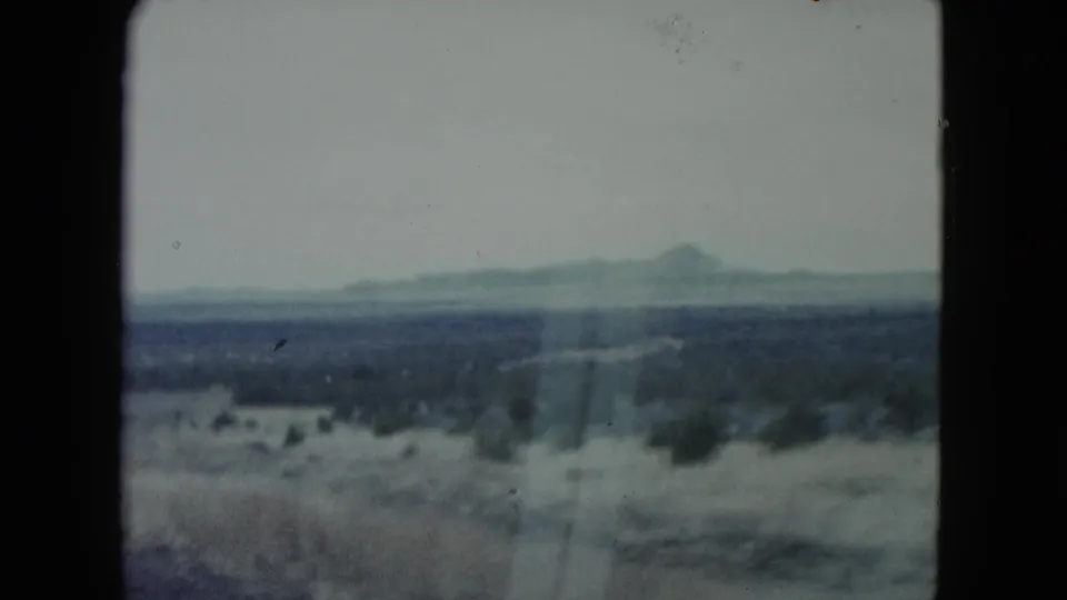 1960 Nevada desert landscape from a moving vehicle, with sparse shrubs, pale winter ground, a faint roadside path, and hazy hills beyond.
