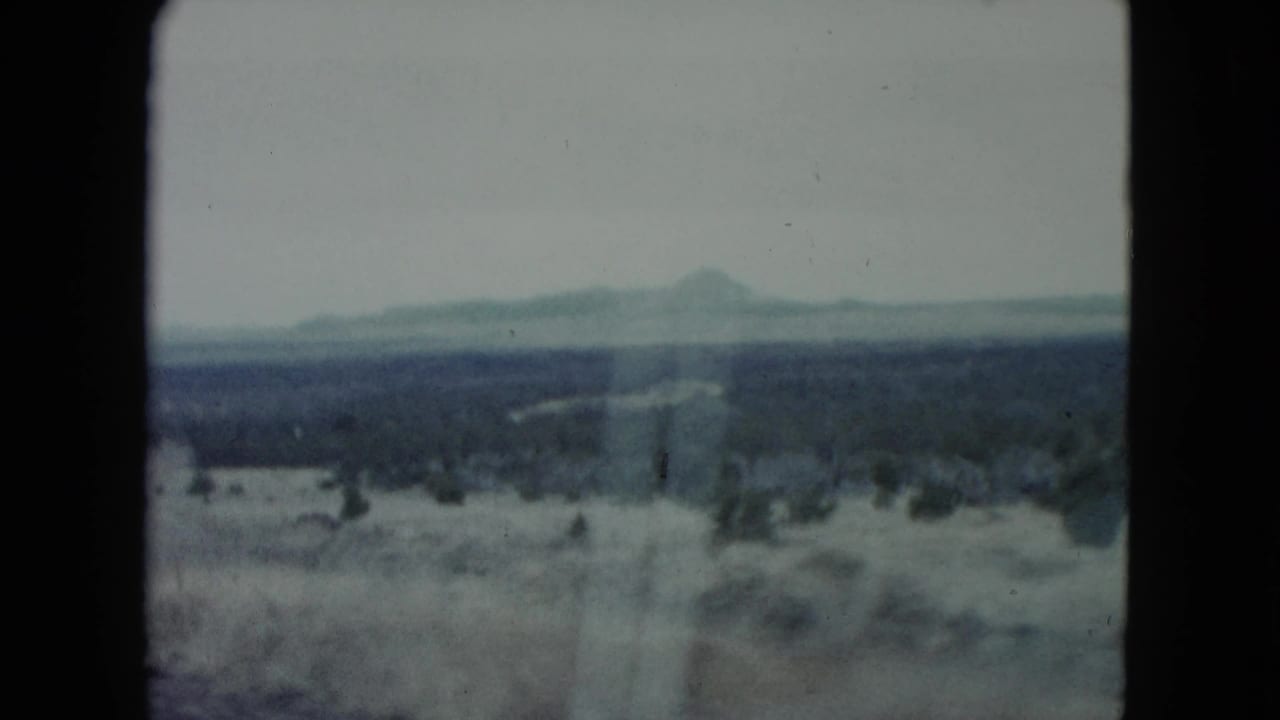 1960 Nevada home-movie frame looking over dry scrubland and distant low mountains from a moving road-trip viewpoint.