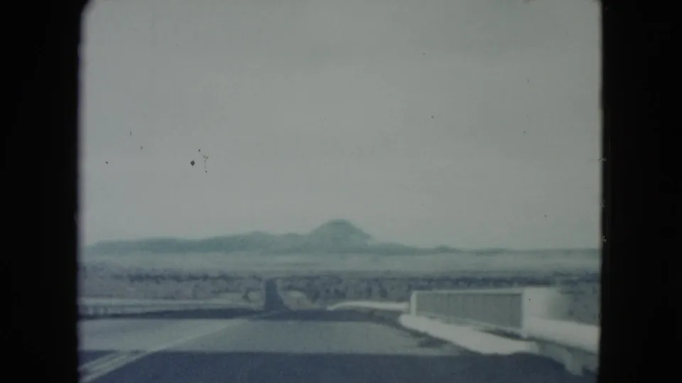 1960 Nevada 8mm frame from a moving car, with paved roadway, bridge rail, dry land, and distant hills ahead.
