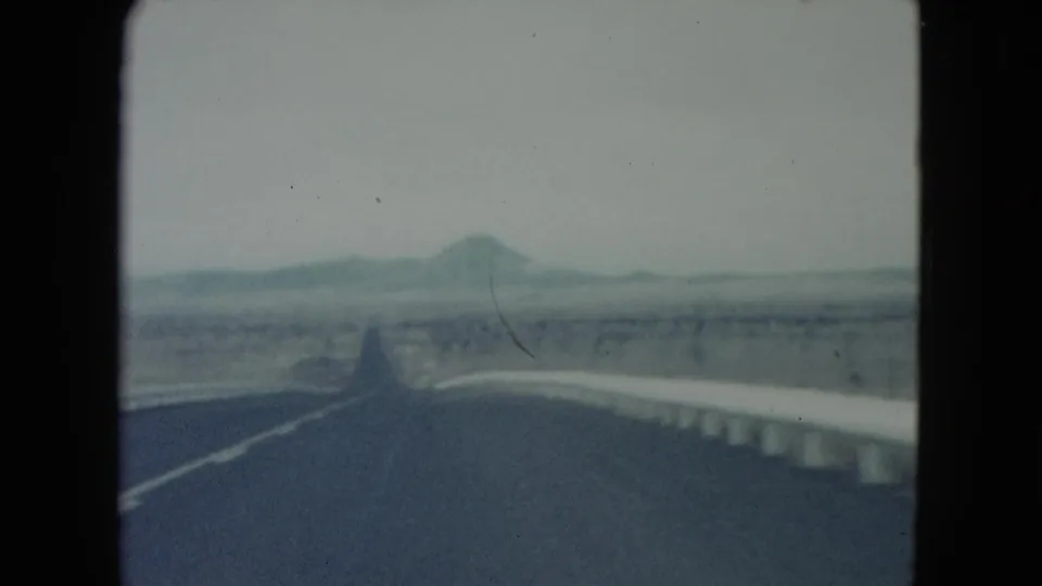 Forward-facing 1960 Nevada road-trip frame showing a paved highway, bridge rails, desert plain, and distant mountains.