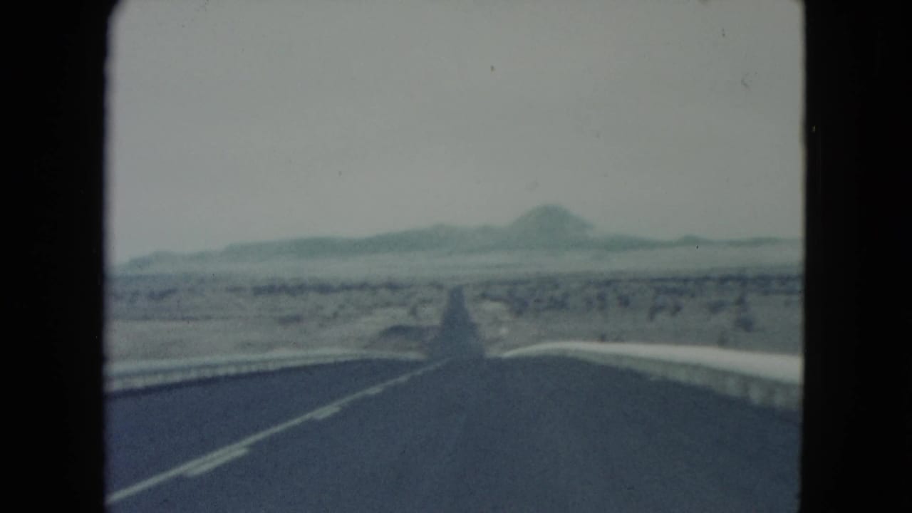 1960 Nevada 8mm frame looking down a straight desert highway toward distant low mountains under a pale winter sky.