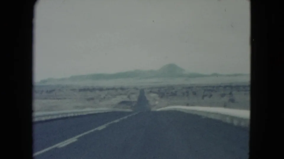1960 Nevada 8mm frame looking down a straight desert highway toward distant low mountains under a pale winter sky.