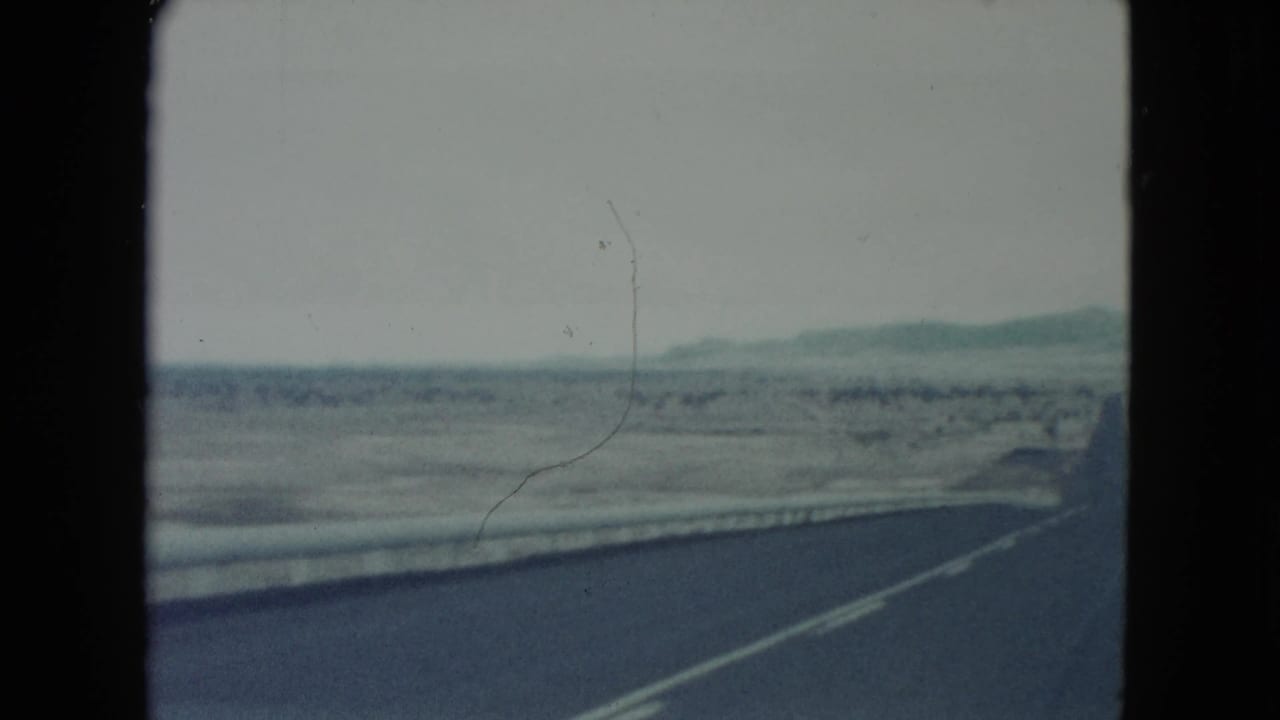 Curving road edge and concrete barrier cross an open desert landscape with distant hills.