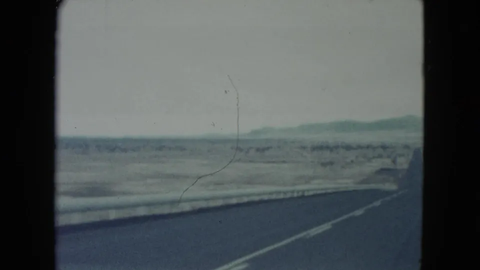 1960 Nevada 8mm frame showing a road edge and barrier passing through open desert with low hills in the distance.
