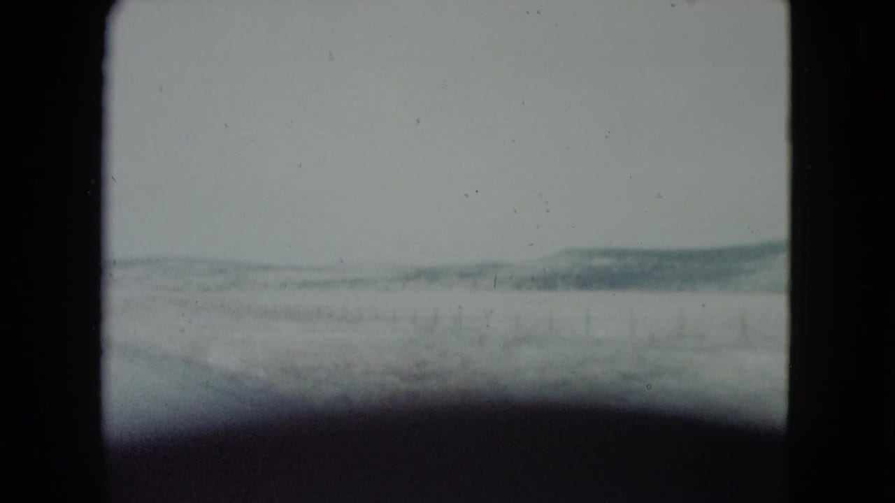 Pale desert landscape seen through a vehicle window with low hills in the distance.
