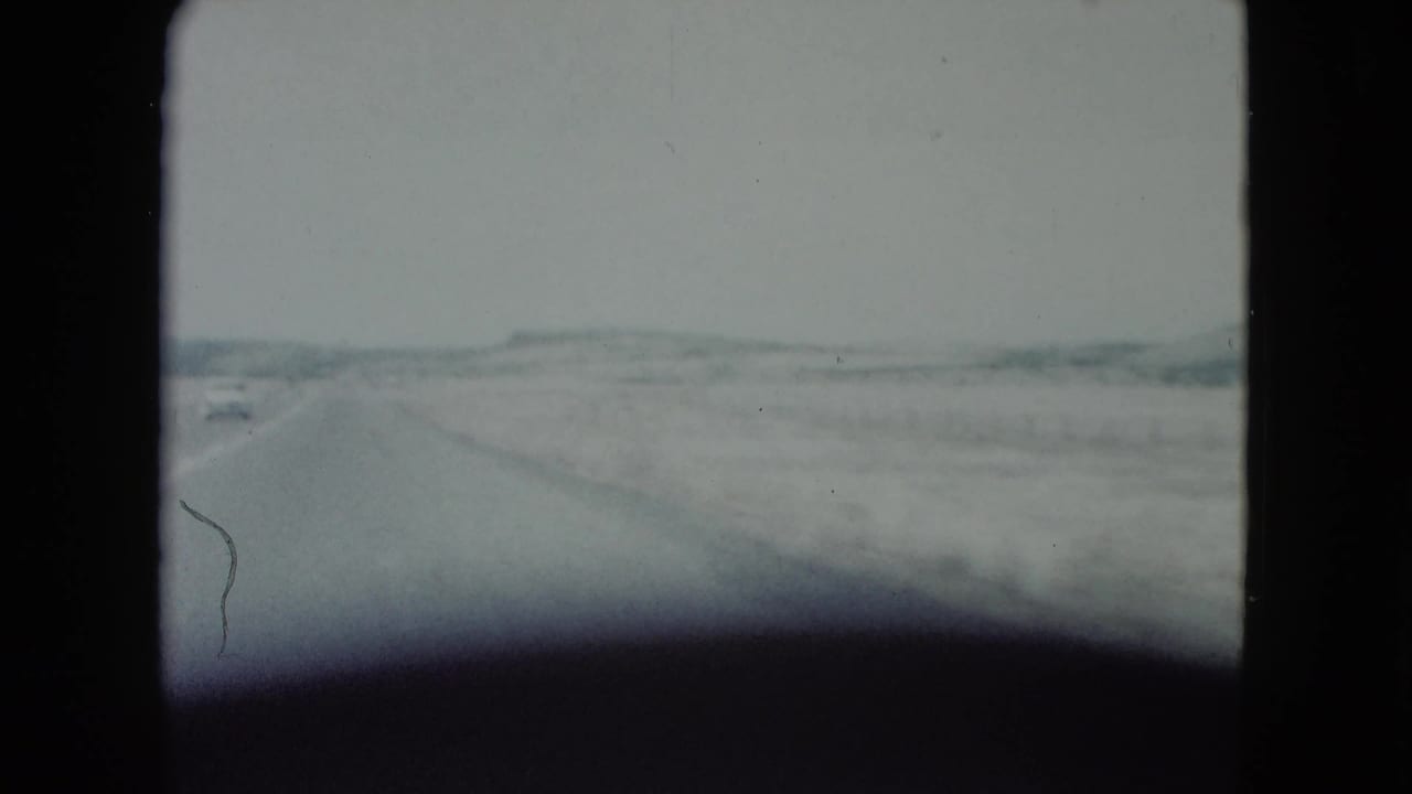 A 1960 Nevada road-trip frame shows a straight desert road, a distant car, and low hills under a pale sky.
