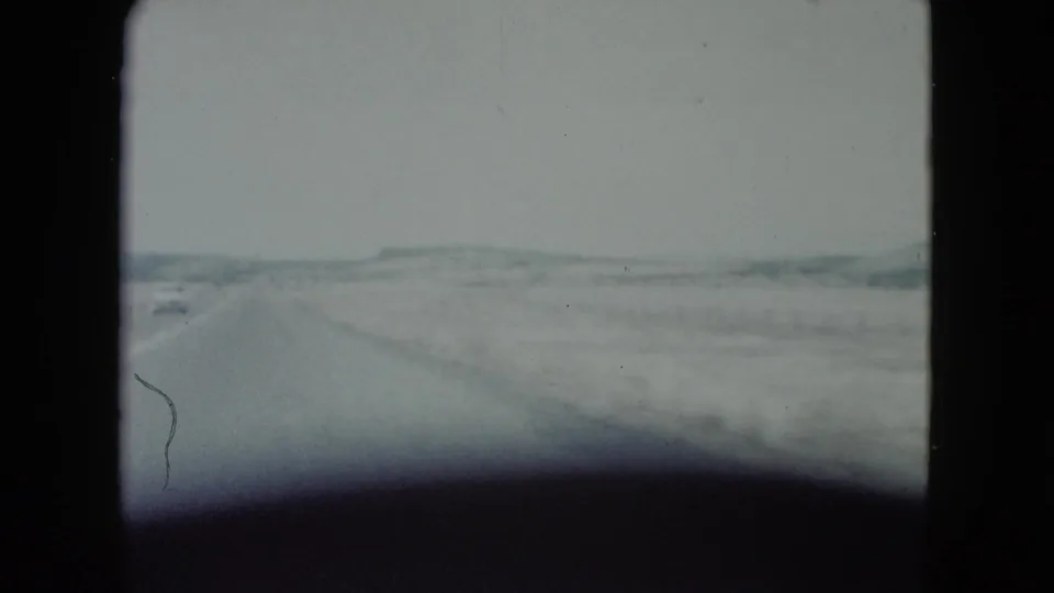 A 1960 Nevada road-trip frame shows a straight desert road, a distant car, and low hills under a pale sky.