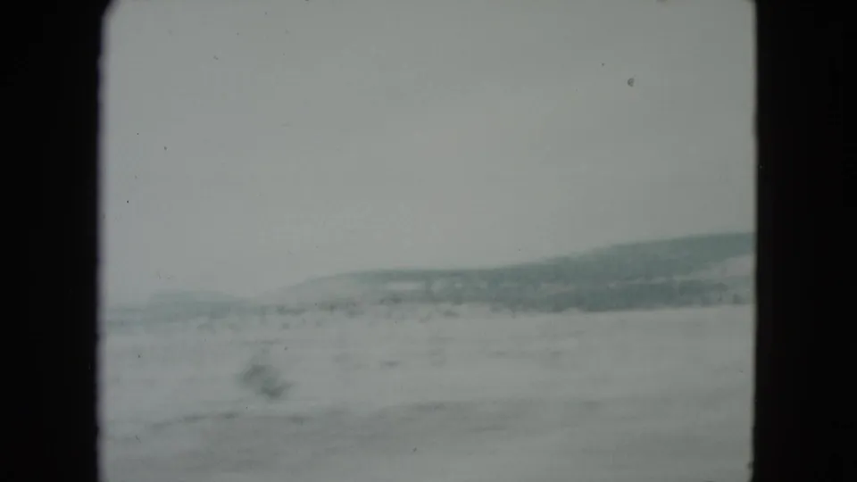 A 1960 Nevada home-movie frame shows open arid terrain and distant hills from inside a moving vehicle.