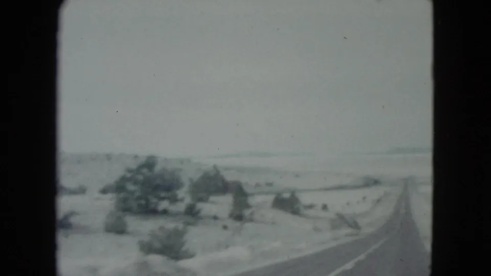 1960 Nevada highway view from a moving vehicle, showing an empty road, sparse desert vegetation, and distant roadside structures.