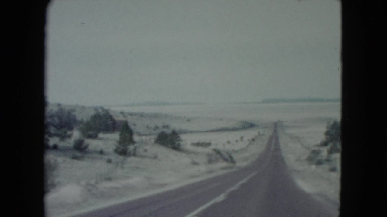 Straight paved road receding through a pale desert landscape with shrubs and low hills.