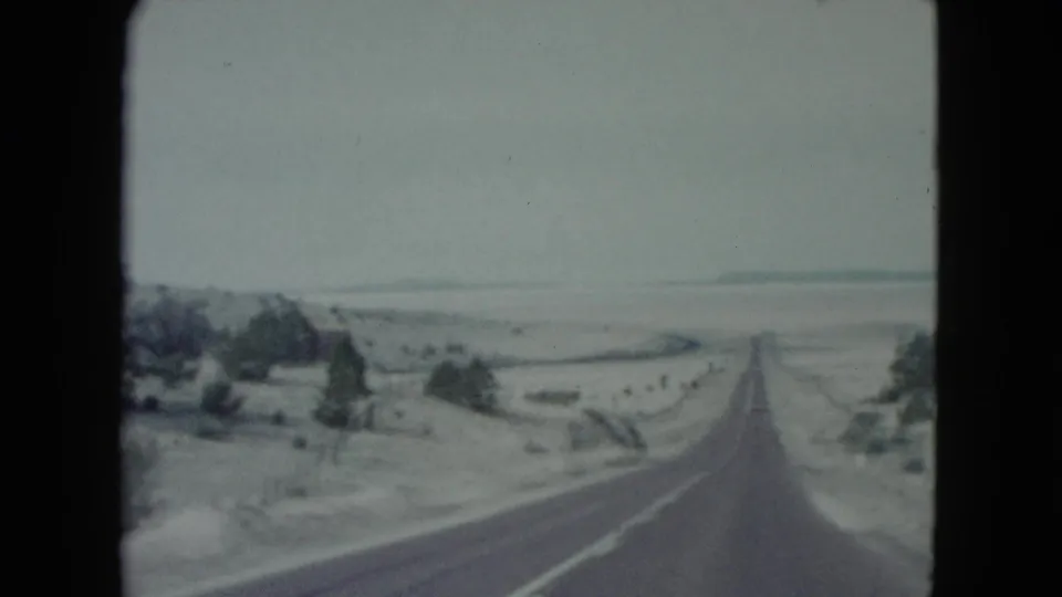 1960 Nevada 8mm frame showing a straight highway stretching through an arid landscape with scattered shrubs, low hills, and muted sky.
