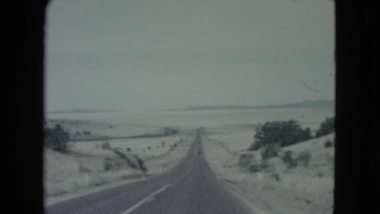 1960 Nevada home-movie frame with a forward view down a two-lane road across open desert terrain and sparse roadside vegetation.