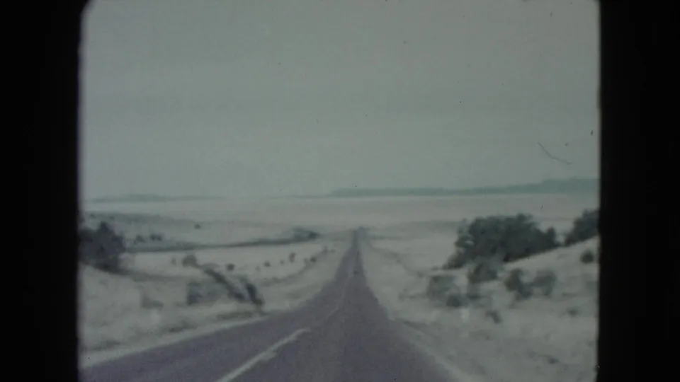 1960 Nevada home-movie frame with a forward view down a two-lane road across open desert terrain and sparse roadside vegetation.