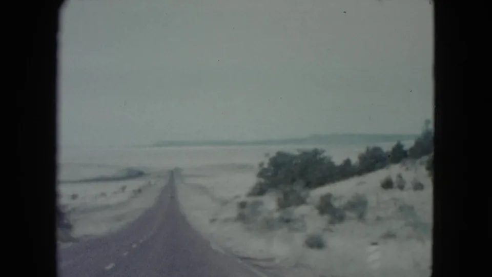 1960 Nevada 8mm frame showing the highway descending through pale desert ground with thicker shrubs and trees along the right side.