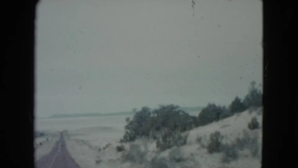 1960 Nevada home-movie frame with the road partly left of center, bordered by a sandy hillside, shrubs, and small trees.