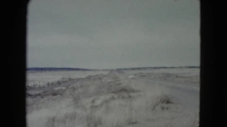 1960 Nevada home-movie frame showing a quiet road bordered by sandy ground and sparse brush under an overcast sky.