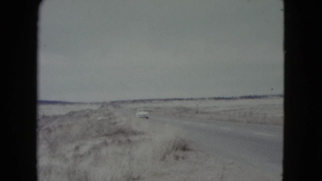 A distant car appears on a rural Nevada road, with dry brush and a low horizon visible in the 8mm frame.