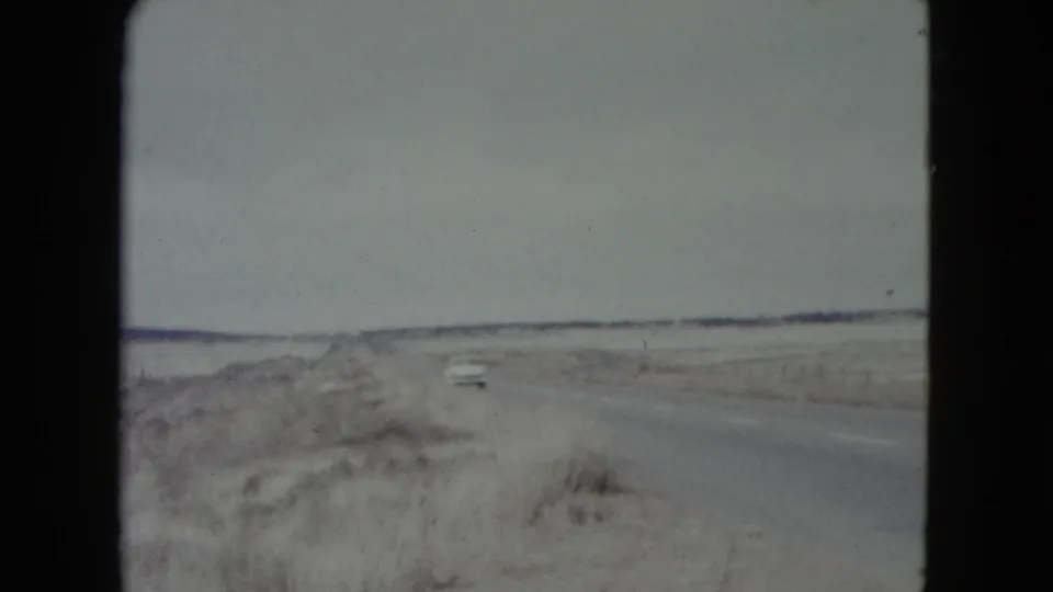 A distant car appears on a rural Nevada road, with dry brush and a low horizon visible in the 8mm frame.