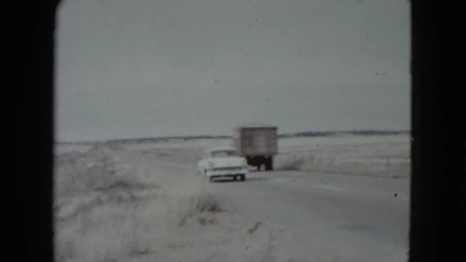 A white mid-century station wagon passes near a box truck on a rural Nevada roadway in grainy 8mm footage.