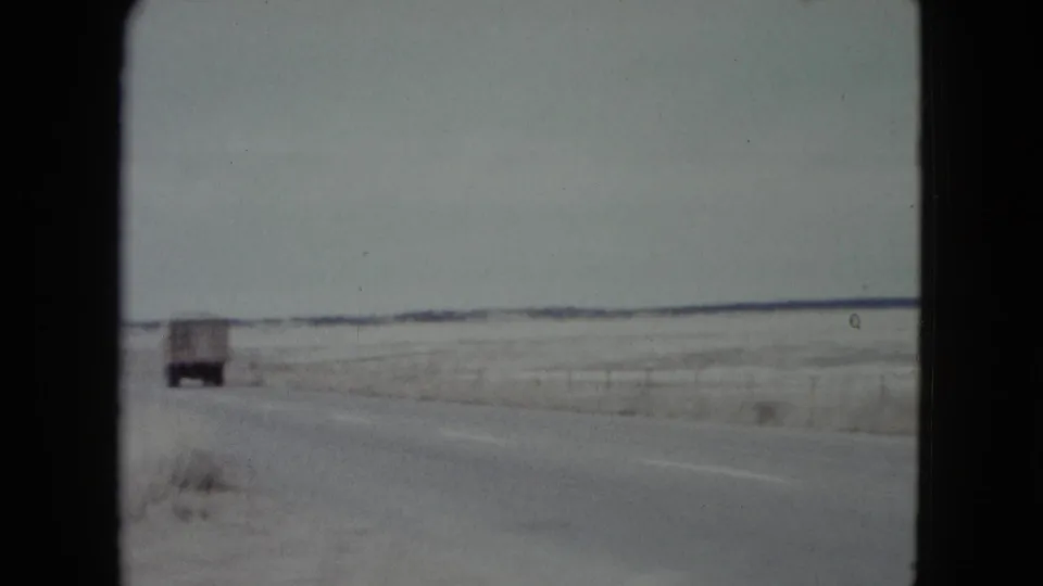 A box truck drives away on a rural Nevada road bordered by sandy ground, dry brush, and a low horizon.
