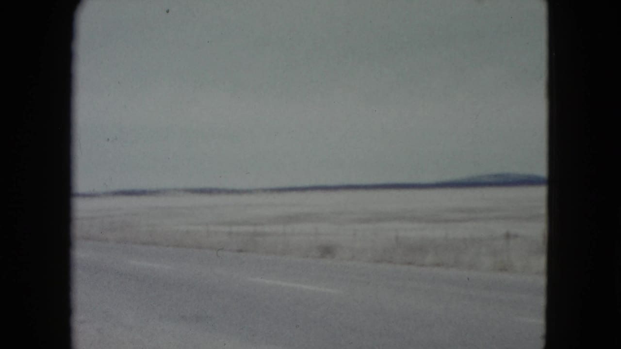 1960 Nevada 8mm frame showing a flat winter plain, roadside edge, muted sky, and low hills on the horizon from a moving vehicle.