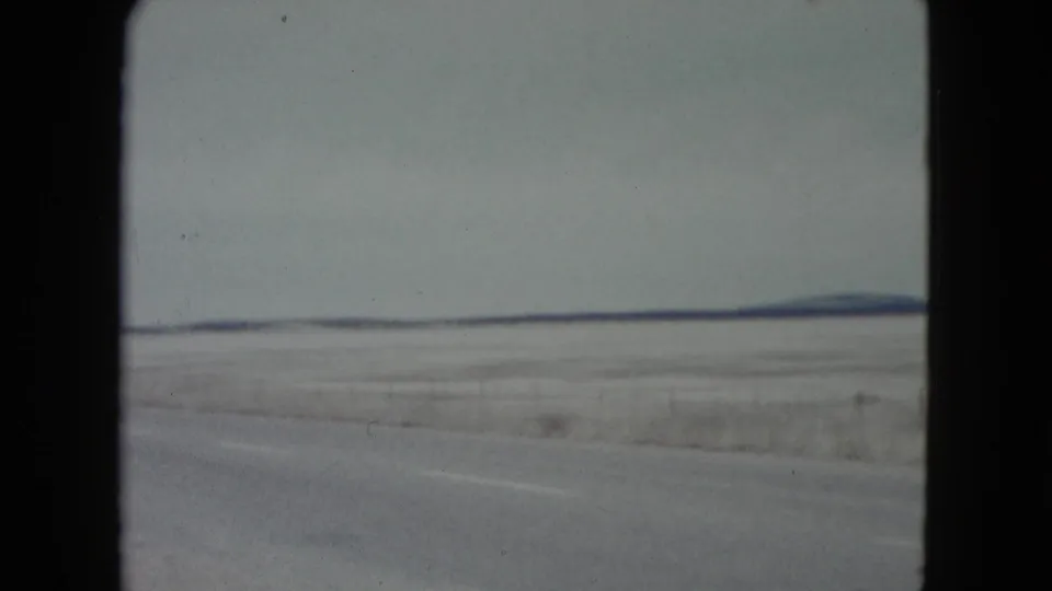 1960 Nevada 8mm frame showing a flat winter plain, roadside edge, muted sky, and low hills on the horizon from a moving vehicle.
