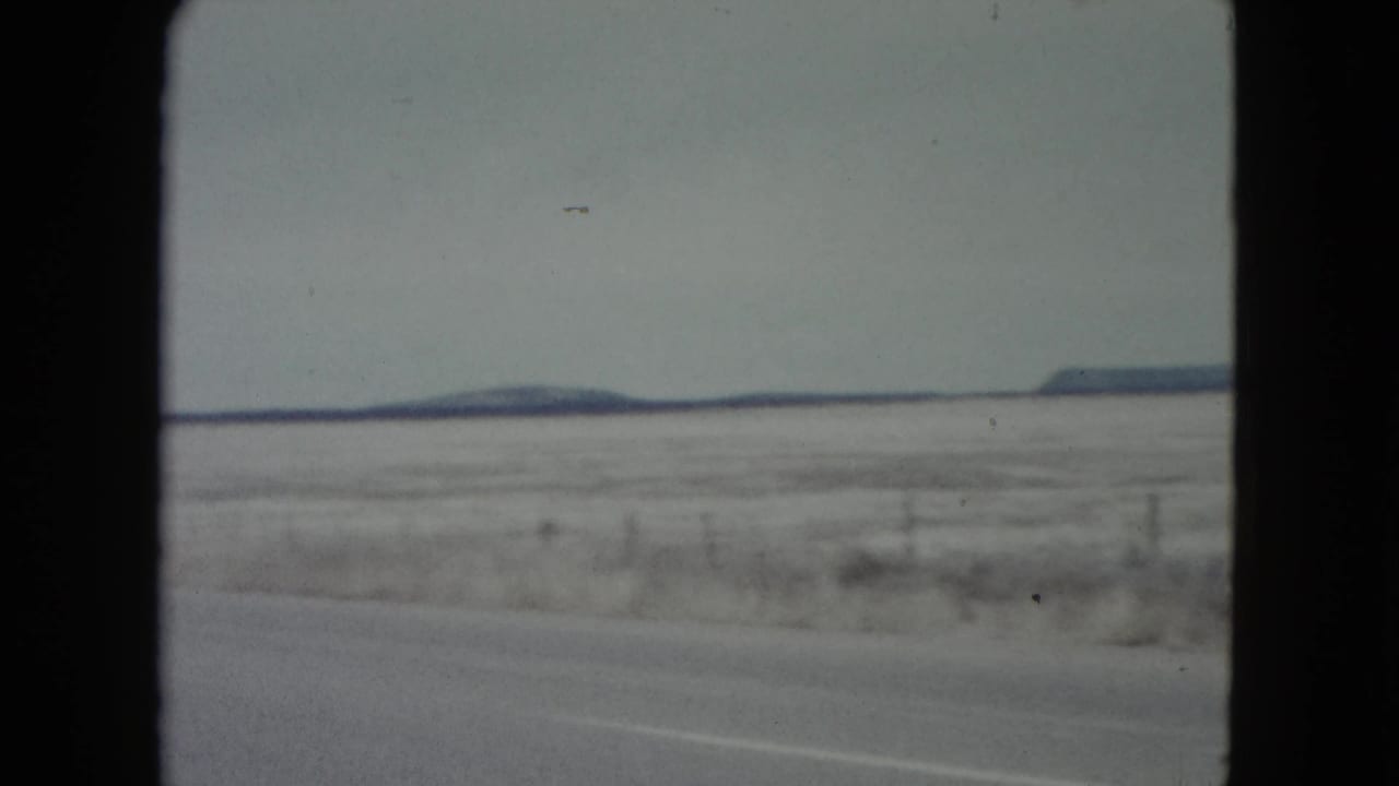 1960 Nevada roadside view with a snow-dusted plain, wire fence posts, a low dark horizon, and soft 8mm motion blur.