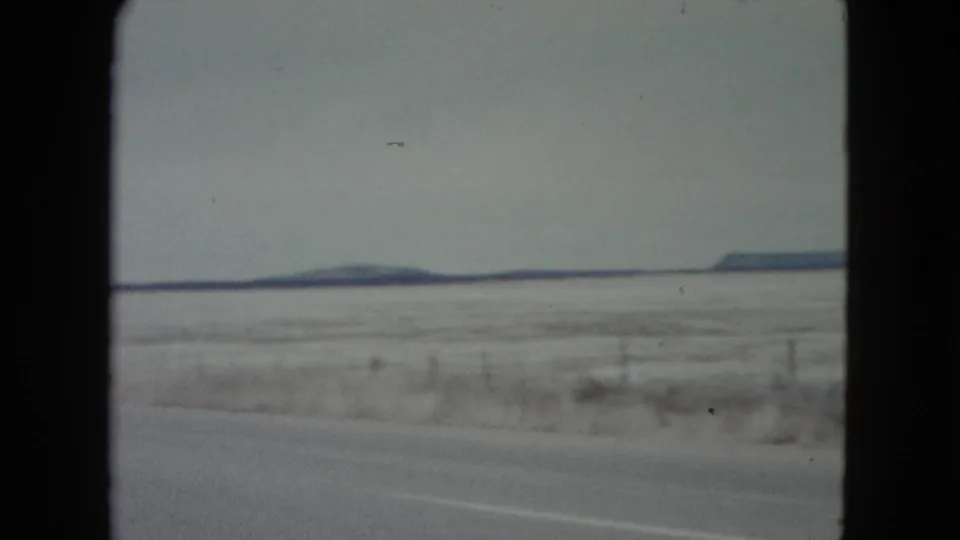 1960 Nevada roadside view with a snow-dusted plain, wire fence posts, a low dark horizon, and soft 8mm motion blur.