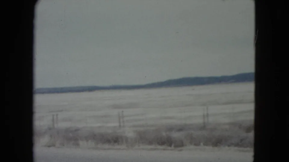 1960 Nevada 8mm frame of a pale open plain, roadside fence posts, and a dark low ridge under a muted sky.