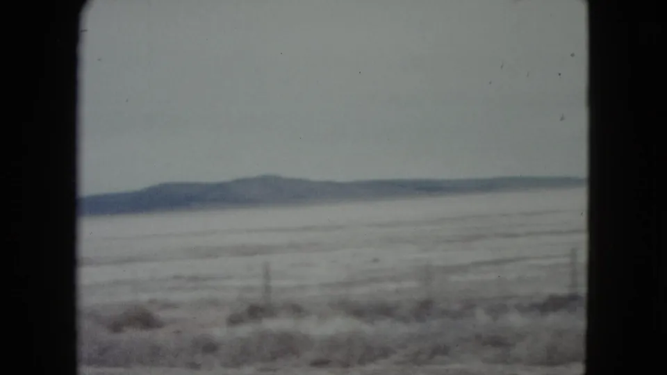 1960 Nevada travel frame showing a snow-covered roadside plain, scattered fence posts, brush, and low hills on the horizon.