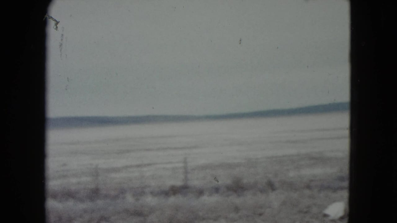 1960 Nevada 8mm frame showing a hazy frozen plain, sparse fence line, and distant low mountains beyond scrubby foreground.