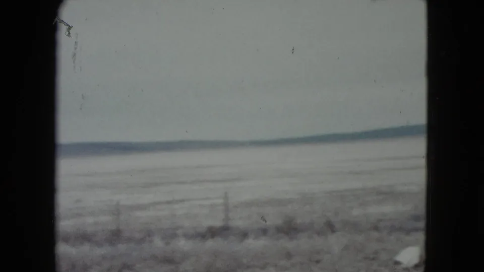 1960 Nevada 8mm frame showing a hazy frozen plain, sparse fence line, and distant low mountains beyond scrubby foreground.