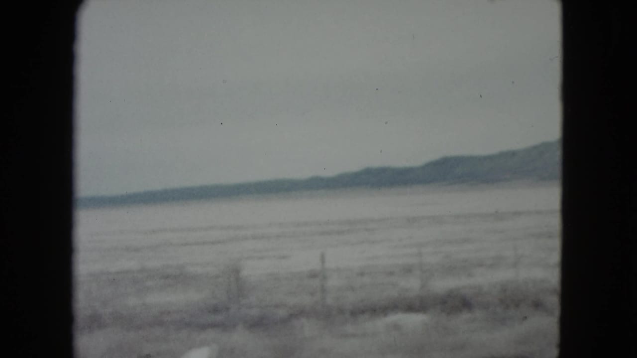 Hazy 1960 Nevada landscape frame with an open frozen plain, low mountain ridge, faint fence posts, and visible 8mm gate borders.