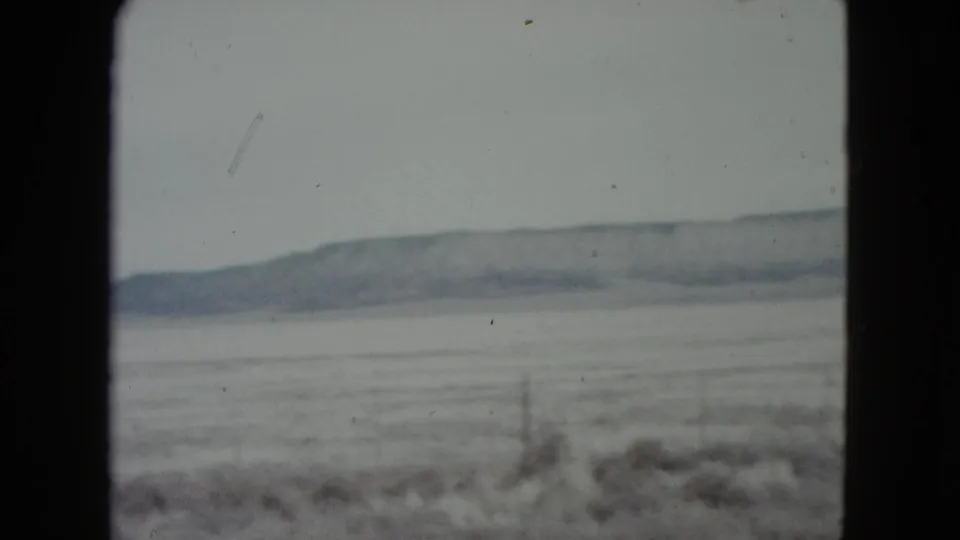 Soft 8mm view of a Nevada frozen plain with scrub, a wire fence, and a low mountain range under a pale hazy sky.
