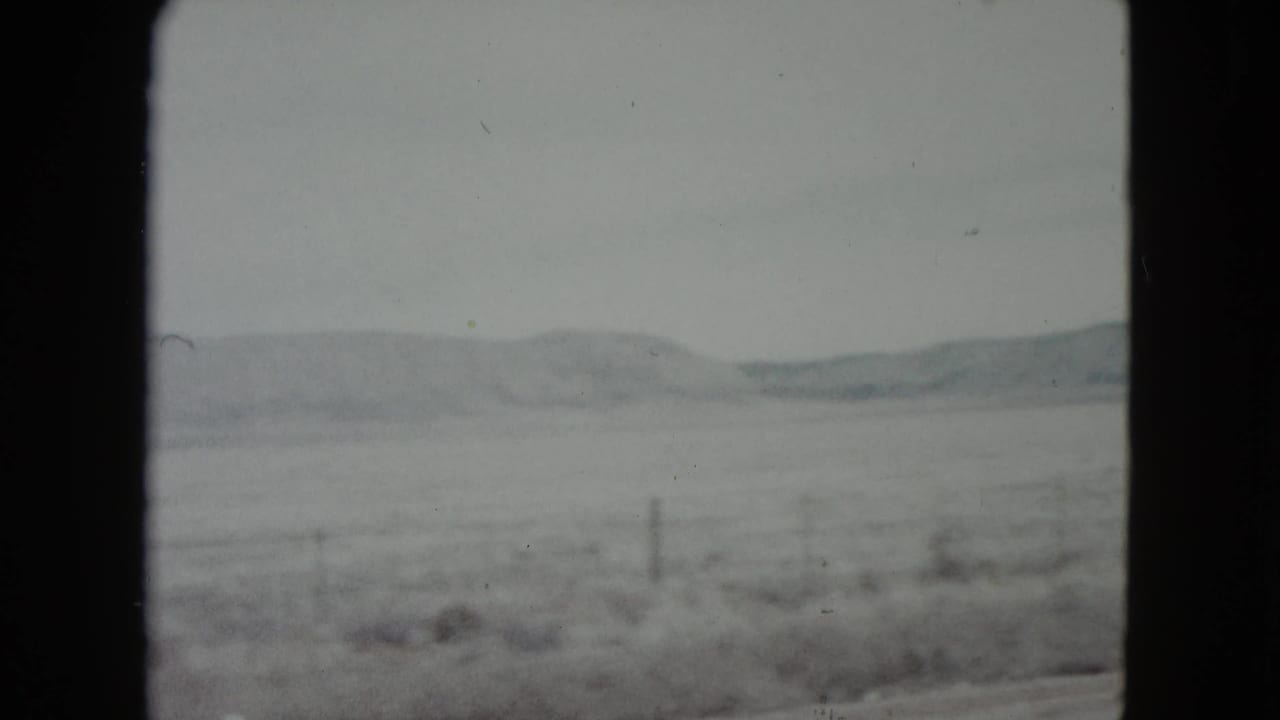 Snow-dusted open field and distant hills seen through a dark vehicle window frame.