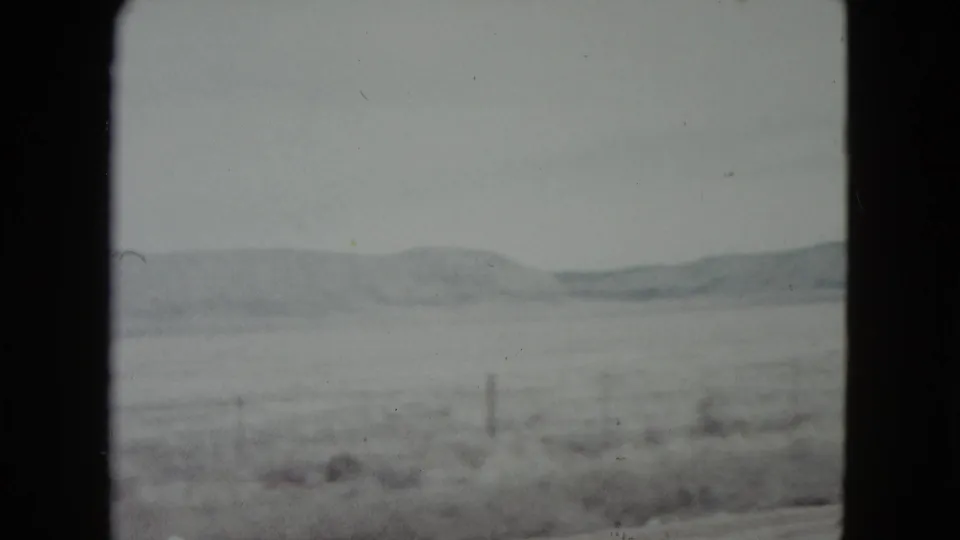 1960 Nevada home-movie frame showing a pale winter field, low hills, and fence posts viewed from inside a moving vehicle.
