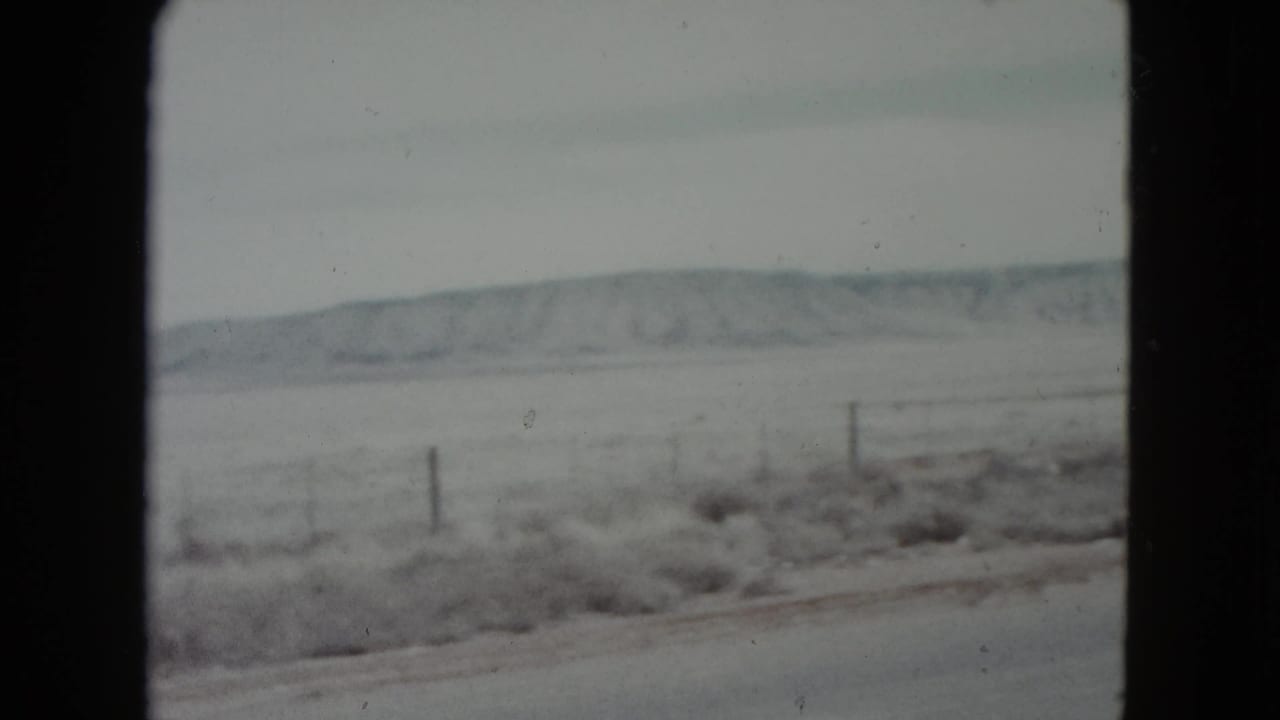 1960 Nevada roadside view with a snow-dusted field, wire fence posts, and distant hills seen from a moving car.