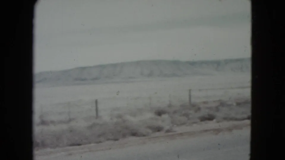 1960 Nevada roadside view with a snow-dusted field, wire fence posts, and distant hills seen from a moving car.