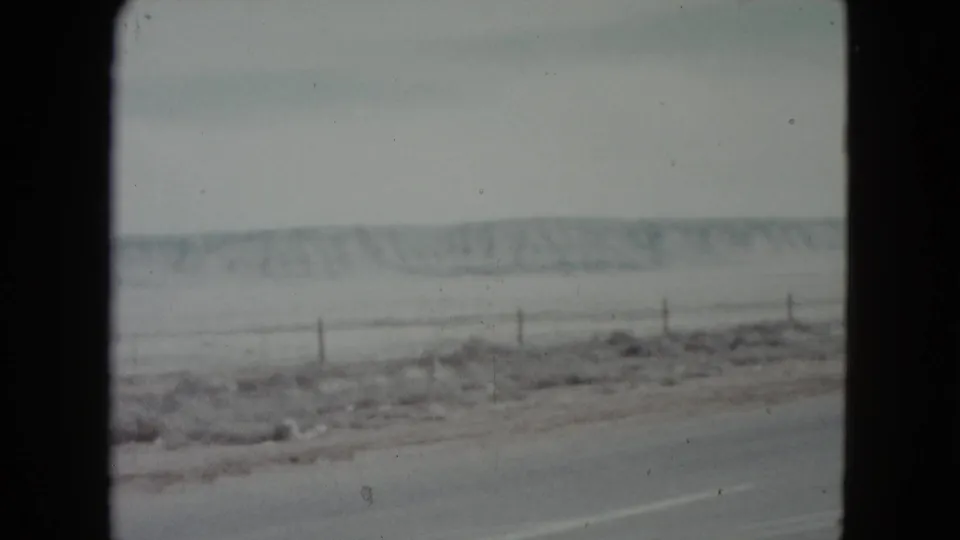 A 1960 Nevada 8mm frame shows a muted winter roadside, fence posts, open field, and low hills from a moving vehicle.