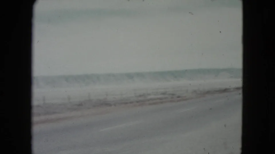 1960 Nevada home-movie frame showing a straight desert road, pale sky, and distant low hills from a moving car.