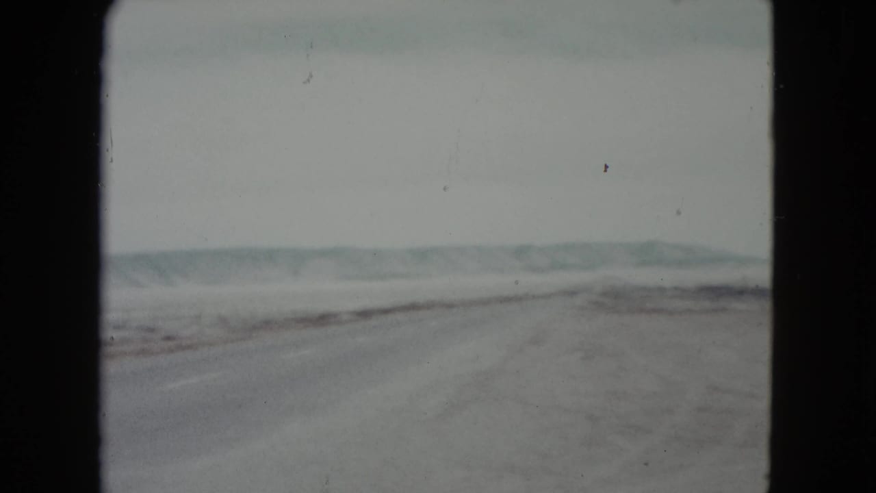 1960 Nevada road-trip view of a long highway through open desert, with low hills and roadside fencing faintly visible.