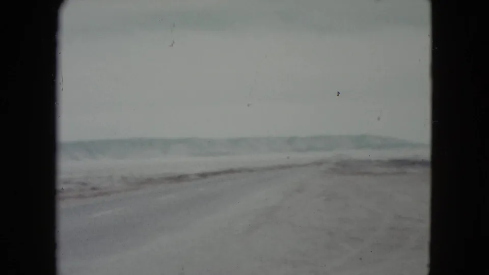 1960 Nevada road-trip view of a long highway through open desert, with low hills and roadside fencing faintly visible.