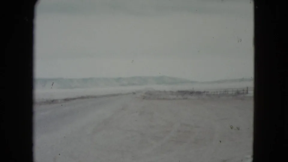 Vintage 8mm view of a Nevada desert road, with scrubby ground, a faint fence line, and low hills beyond the highway.