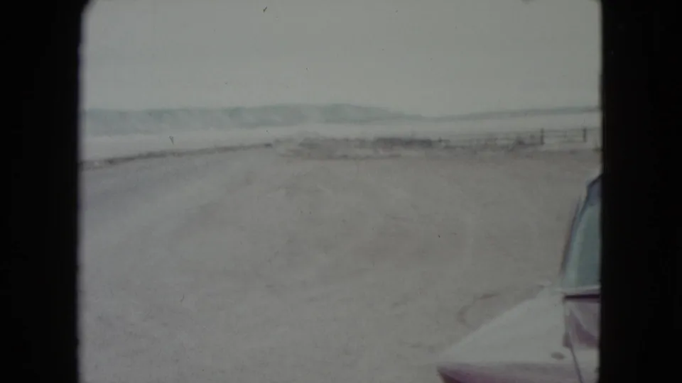 1960 Nevada road-trip frame showing the highway ahead, sparse desert fencing, distant hills, and part of a car at the right edge.