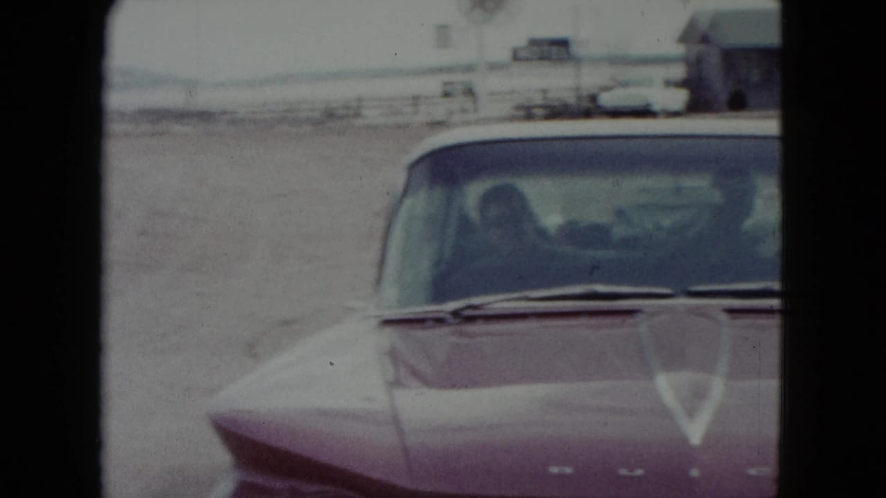1960 Nevada 8mm frame of a red car near a roadside gas station, with a driver or passenger silhouette visible through the windshield.